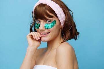 a happy woman stands on a blue background in a soft rim and with colored, shiny eye patches, smiling sweetly holding her hand near her face