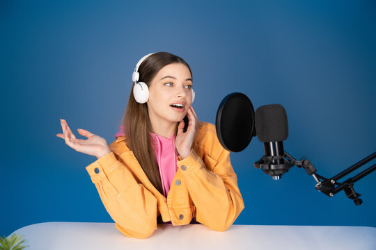 Teen girl in headphones talking during podcast near studio microphone isolated on blue. - Powered by Adobe