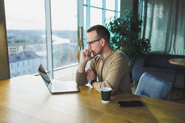 Young freelancer man in glasses sitting at a wooden table with a laptop and coffee while working on a remote project in a modern workspace.