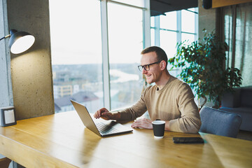 Young freelancer man in glasses sitting at a wooden table with a laptop and coffee while working on a remote project in a modern workspace.