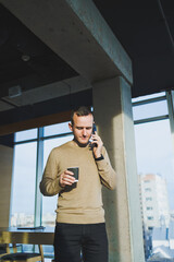 Positive young man smiling and talking on mobile phone and drinking coffee while resting while standing in office. Lunch break during work.