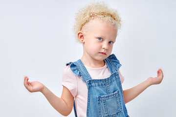 Calm confused pretty european little girl in dress spreads arms to the sides, isolated on white studio background, empty space, studio shot. Big choice, offer and advertisement. copy space