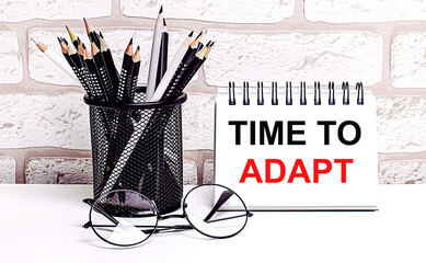 A workplace in the house with glasses, pencils in a stand and a notebook with text TIME TO ADAPT on...