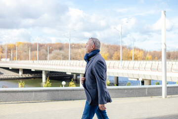 Calm gray haired man looking away camera in warm jacket and scarf walking street, near promenade bridge. Low temperature