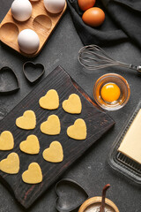 Wooden board with raw heart shaped cookies, ingredients and cutters on dark background. Valentines Day celebration