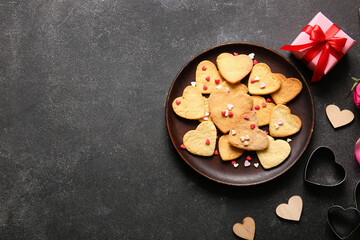 Composition with plate of tasty heart shaped cookies, cutters and gift on dark background. Valentines Day celebration