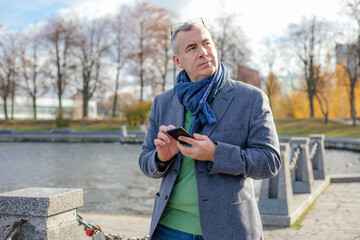 Obraz premium Thoughtful, busy, modern nervous grizzled man use smartphone and looking away while walking in park near river outdoors