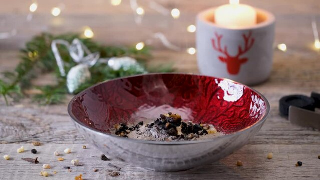 Mixture of aromatic incense resin, frankincense (olibanum), styrax and rose blossom petals burning on hot charcoal in a red incense bowl on a wooden table with christmas decoration and candle, closeup