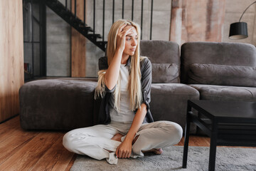 Obraz premium Tired blonde beautiful woman sitting on floor at home, touching forehead, Swedish housewife exhausted after hard work. Overloaded pretty female relaxing. Upset beautiful lady after divorce, headache.