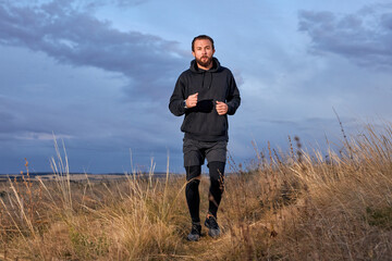 Naklejka na ściany i meble Jogger Young Caucasian Male Running Through the field. Man Wearing Black sportive wear. athlete guy with beard is motivated to be muscular, strong, in perfect physical shape. sport, fitness