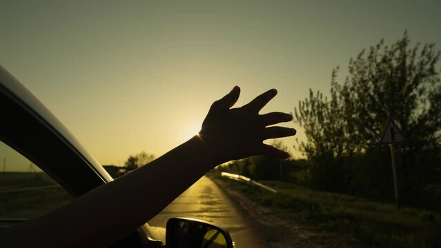 Free woman driver rides car catches wind with her hand from car window. Girl with long hair sits in front seat of car, her hand out window and catching wind, glare of setting sun. Child travels by car