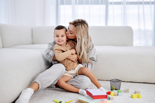 Portrait of 30s woman kissing small cute child boy, sitting on warm floor at home. Happy young mother, preschool kid son is looking at camera, posing for family photo. family, children,love concept