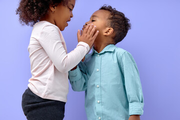 Two cheerful black american siblings, sister kissing her brother, isolated on purple background. portrait of cute kids in casual clothes. Boy and girl feeling love to each other.