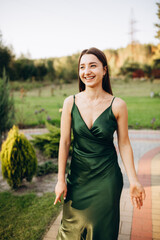 Beautiful smiling girl in a dark green dress with straps. Cute model girl stands on green grass and smiles. Brunette in the summer at a party. Emerald satin dress