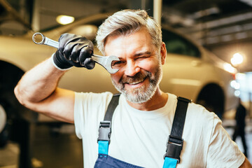 Portrait of pleased proffesional grey haired american car technician in the workplace. Man mechanic in overalls holding wrench in garage.
