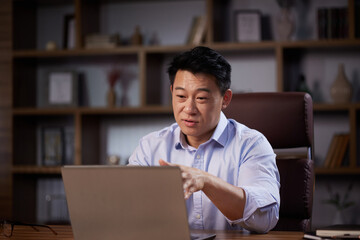 Asian man holding online webinar using laptop webcam, sitting in a home office. Man having online meeting and communicating remotely, looking at computer screen