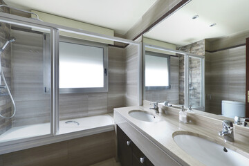 Corner of a toilet with a large mirror on the wall, a two-bowl sink in white porcelain and beige marble and a hydromassage unit with a glass partition