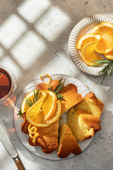 Sliced fluffy orange cake decorated with fresh orange slices with tea and window shadow on light gray textured background, sunny morning breakfast in harsh light. Text space, flat lay, top view
