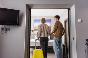 back view of young tourists with suitcases holding hands in modern hotel suite.