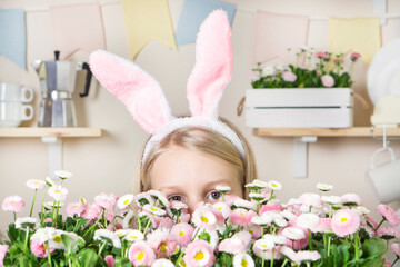 Obraz na płótnie Canvas Easter A little girl with rabbit ears peeks out or hides behind daisy flowers against the backdrop of a home kitchen decorated for Easter. copy space