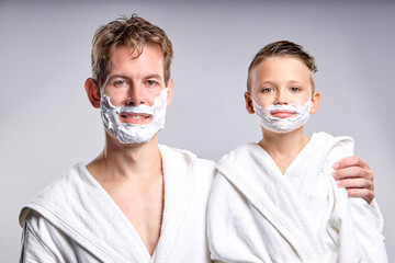 Friendly confident young father and teenage son with shaving foam on faces are looking at camera, wearing white bathrobe, at weekends, posing together, daily routine, family relationships