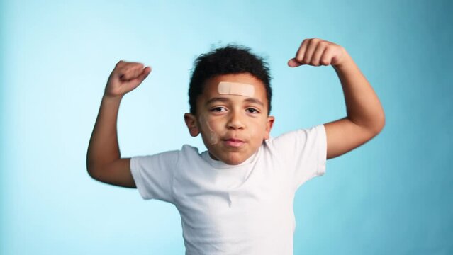 Portrait of clumsy brave smiling african american little boy with adhesive plaster looking at the camera and showing his muscles on isolated blue background Happy childhood 