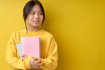 Pretty cheerful attractive beautiful young brunette asian woman 20s wearing basic casual yellow shirt standing with books, looking at side, isolated on bright yellow colour background, in studio