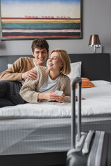 happy woman laughing and holding cellphone near young boyfriend in hotel bedroom on blurred foreground.