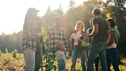 Group of males and females volunteers with kid standing in field of forest and listenig to curator before planting trees. Seedlings in hands. Men and women plant park. Ecological environment concept.