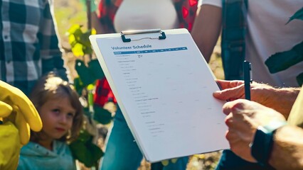 Close up of volunteer schedule in hands of man, curator of eco gardening project. Outdoor. Kid helping to adults in volunteering. Giving orders and sharing tasks. Environmental cooperation.