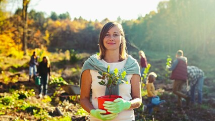Portrait shot of young beautiful Caucasian woman standing outside with tree seedling in pot and...