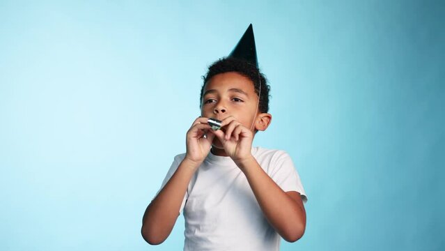 Cute happy little african american boy kid with birthday hat blowing party trumpet during celebration on isolated blue background Adorable child having fun during his birthday at studio