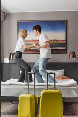Fototapeta na wymiar side view of excited couple holding hands and having fun on bed near baggage in modern hotel bedroom.