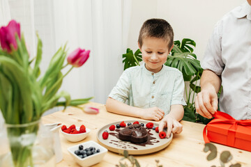 Dad and son decorate a heart-shaped cake with fresh berries and prepare a gift. Mother's Day, International Women's Day, Valentine's Day