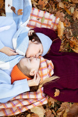 Obraz na płótnie Canvas Vertical glad, satisfied, pretty young mother kiss gently her little daughter in warm clothes and hats lying on ground
