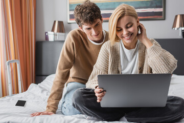 smiling blonde woman sitting with laptop on hotel bed near young boyfriend.