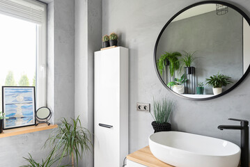 Stylish bathroom with ceramic washbasin, round mirror on the grey wall and window with picture....