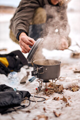 cropped unrecognizable man preparing food in winter forest. Bushcraft, travel, adventure, tourism and people concept. smoke from prepared cooked meal on small saucepan.
