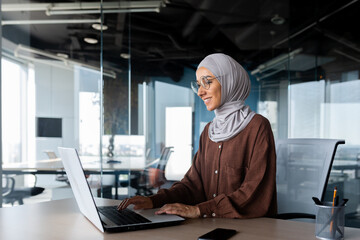 Young beautiful muslim woman in hijab working inside modern office, businesswoman using laptop at...