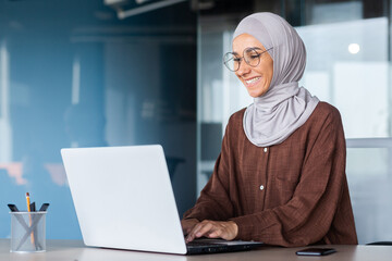 Young beautiful muslim woman in hijab working inside modern office, businesswoman using laptop at work, typing on keyboard while sitting at workplace smiling and happy with achievement result.