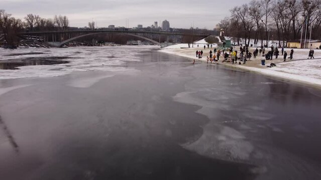 the drone flies over the hole in the winter river, the river is covered with ice, people enter the cold water, cross themselves and plunge headlong into the ice water three times
