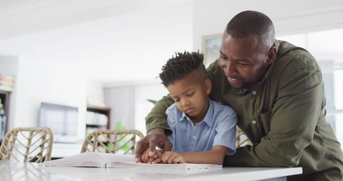 Happy african american man and his son sitting at table and reading braille