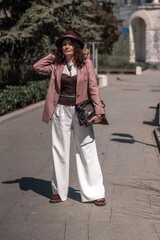 Woman park city. Stylish woman in a hat walks in a park in the city. Dressed in white corset trousers and a pink jacket with a bag in her hands.