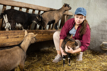 Young female farm worker kneeling to play with baby goats