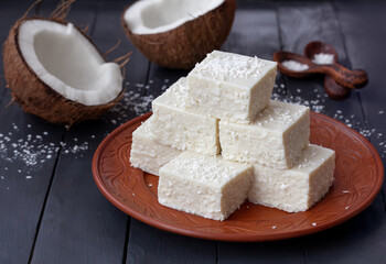 Homemade coconut pudding or white yogurt jelly squares served on a plate with split coconut on background. Dark wooden table, selective focus, horizontal.