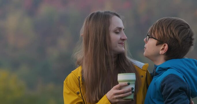 Boy kisses and hugs mother on nature in autumn. Mom laughs and hugs teenage son outside with mug in her hand