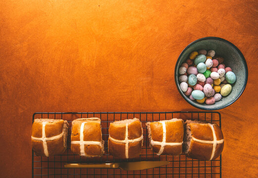 Hot Cross Buns Laid On A Black Wire Rack And A Bowl Of Speckled Easter Eggs. Orange Rusty Copper Coloured Surface Background.