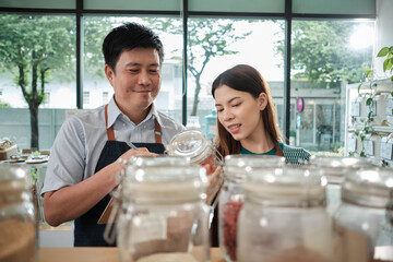 Two shopkeeper colleagues check stock of natural organic products in reusable containers shelves at refill store, zero waste, plastic-free grocery shop, and eco-friendly, sustainable lifestyles.