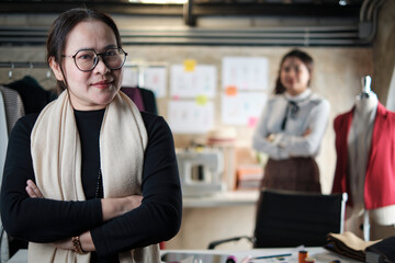 Asian mature female fashion designer looks at camera and smiles in studio, arms crossed, working with creative and sewing for dress design collection, professional boutique tailor SME entrepreneur.