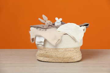 Laundry basket with baby clothes and crochet toys on wooden table against orange background